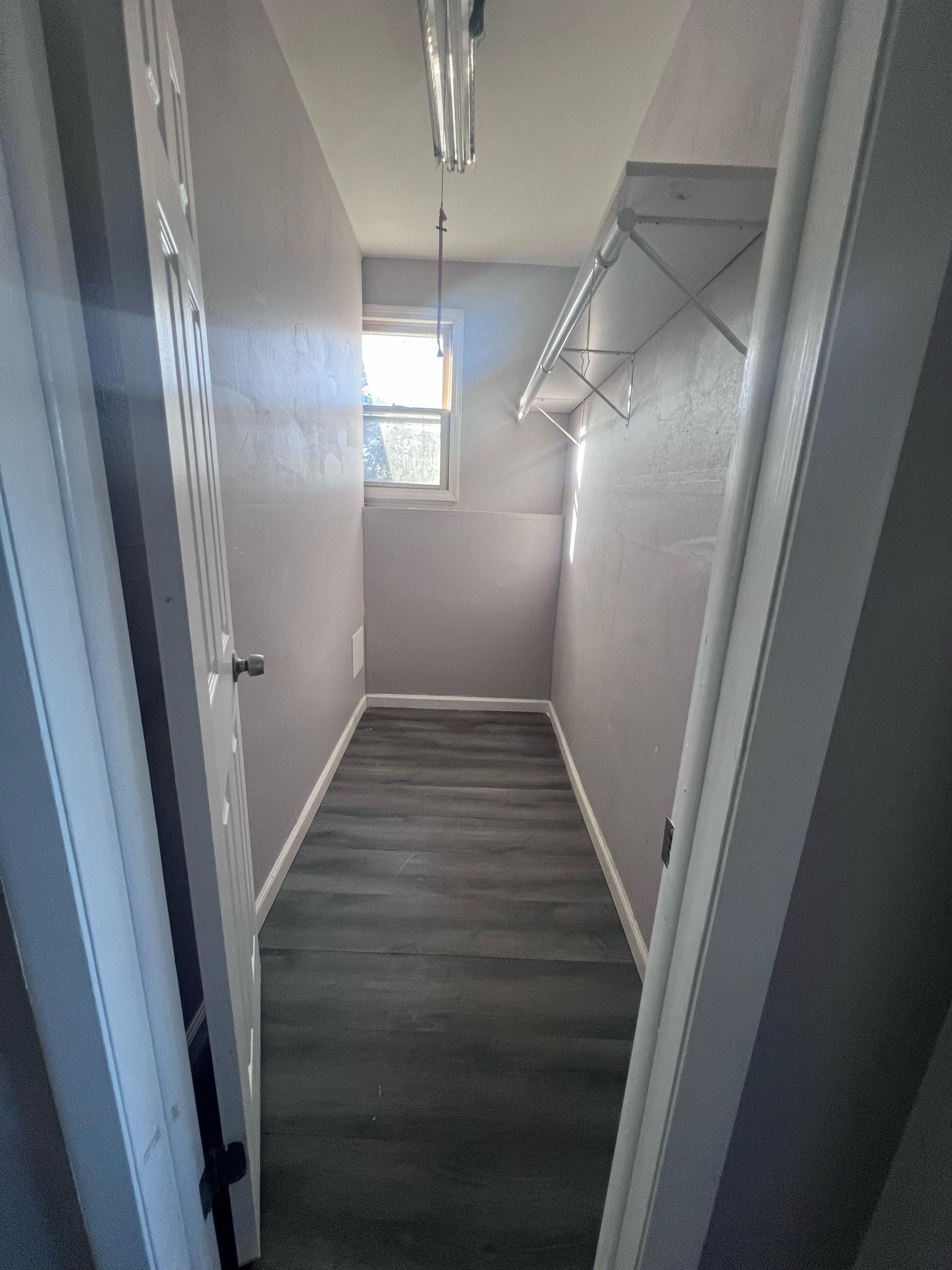 Narrow walk-in closet with gray walls, wood-look flooring, window, and white hanging shelf system.