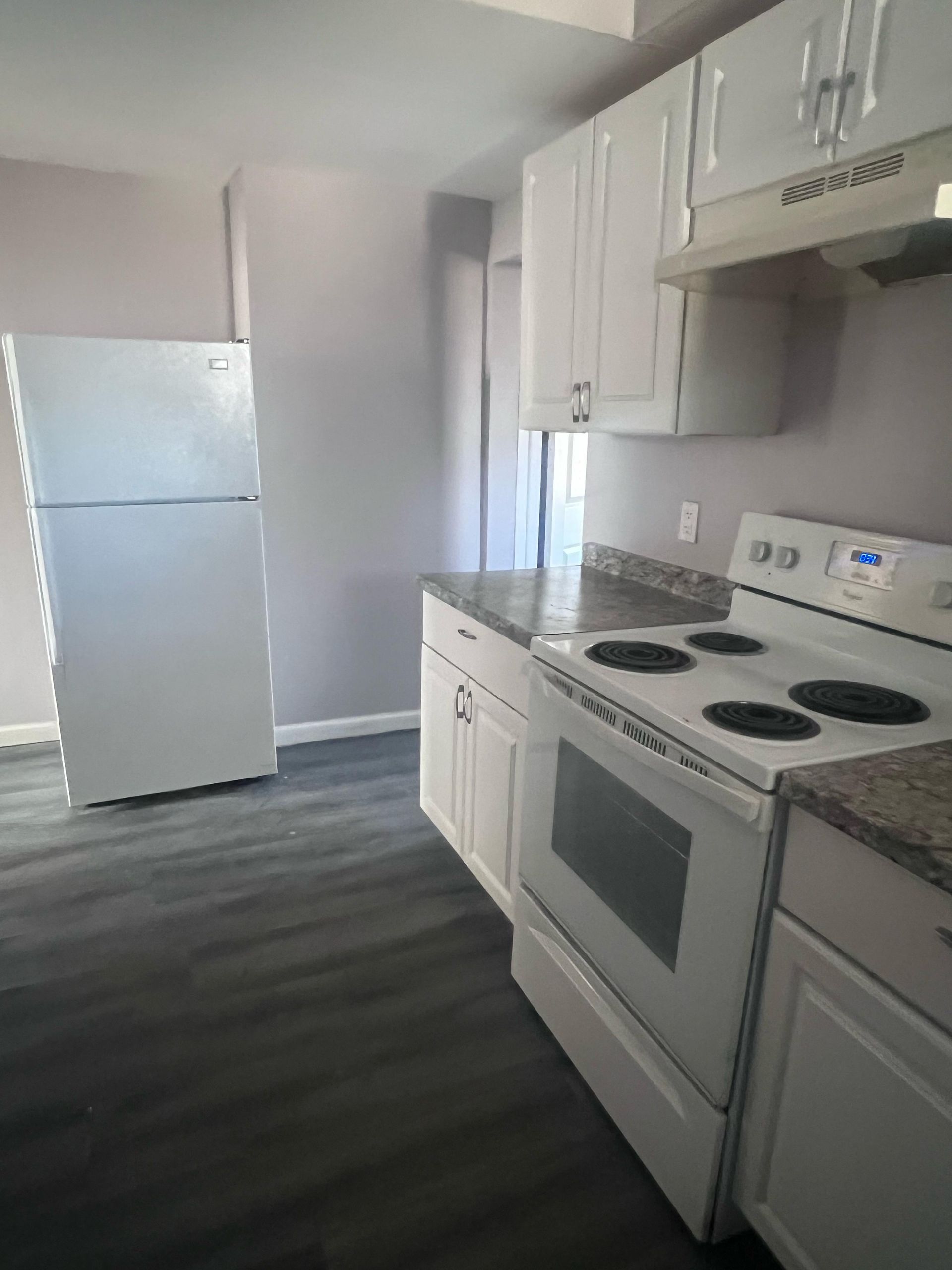 Empty modern kitchen with white cabinets, stove, refrigerator, and gray wood-look flooring.