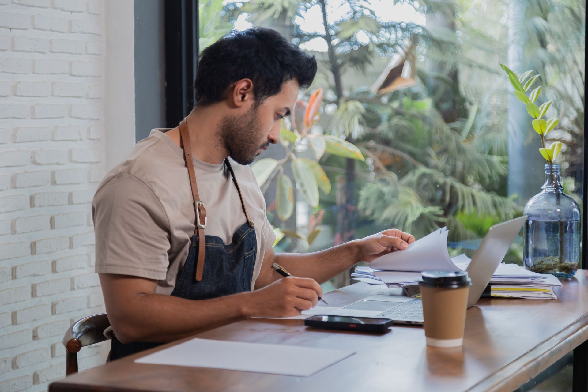Man in apron working at a cafe table, reviewing papers and using a laptop, near a window.