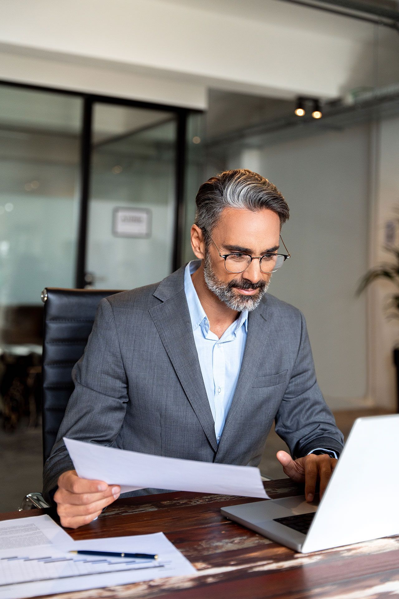Man in gray suit working at a laptop, reviewing paperwork in a modern office.