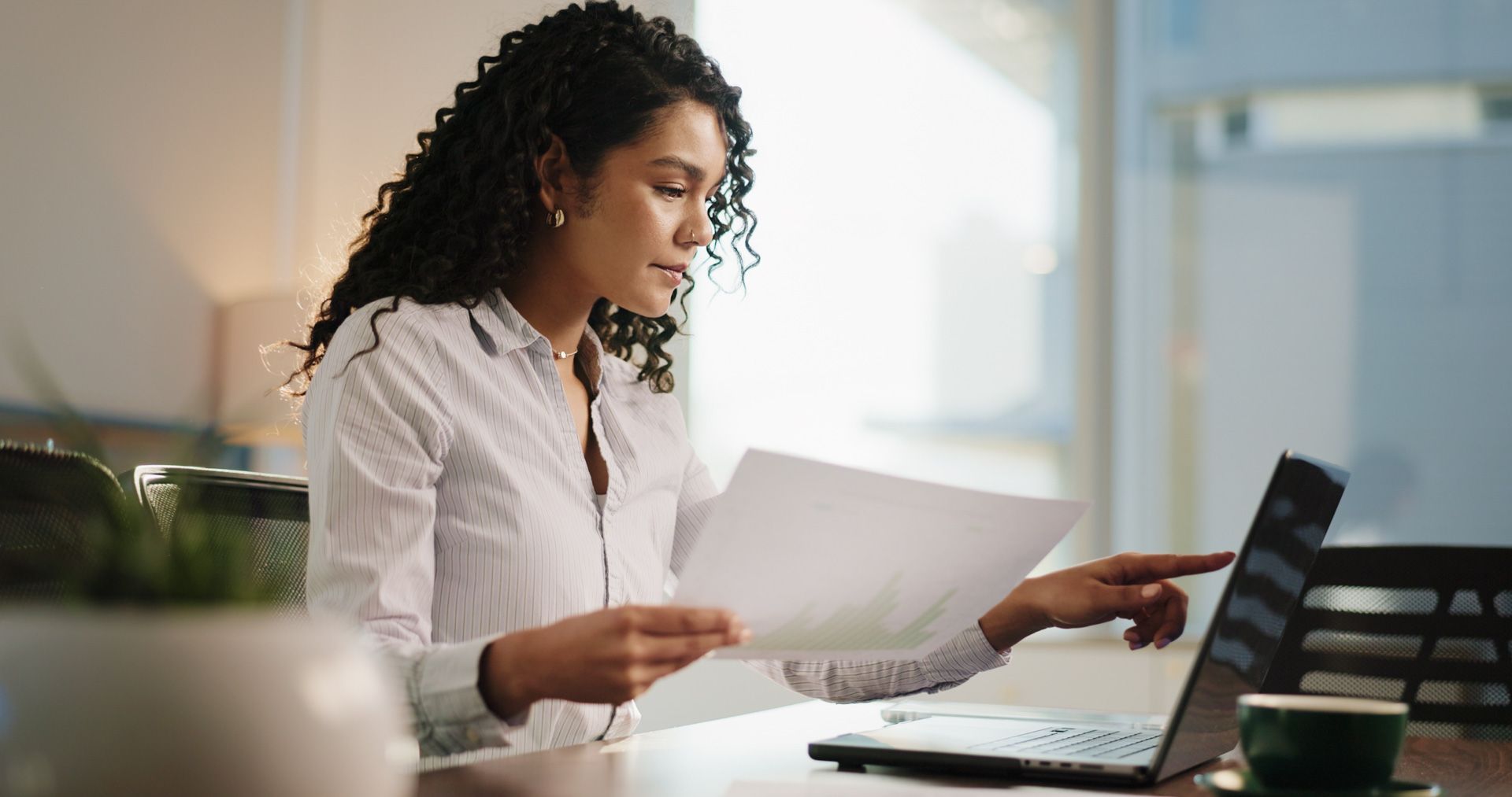 Woman with curly hair works on laptop, holding papers, pointing at the screen at a desk in a well-lit space.