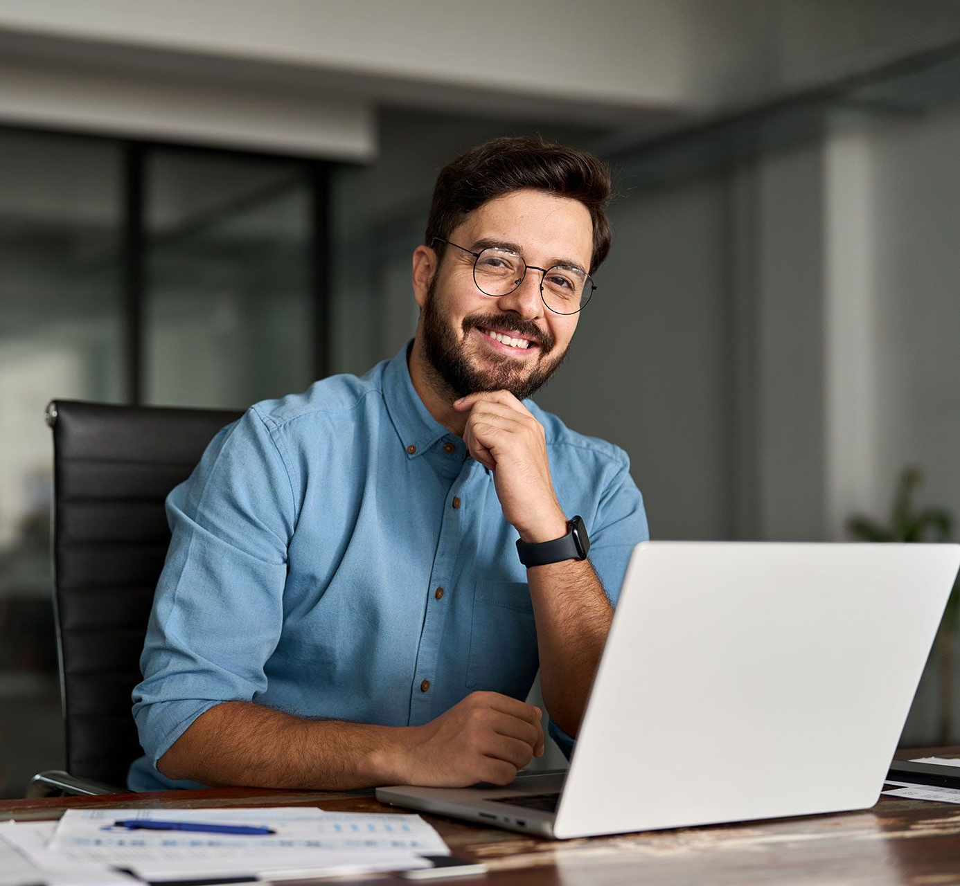 Man with a beard and glasses smiling at the camera, sitting at a desk with a laptop.