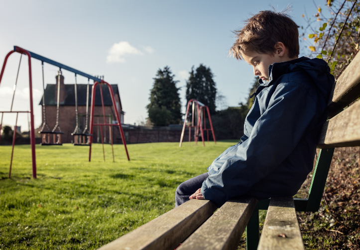 A young boy is sitting on a bench in front of a playground.