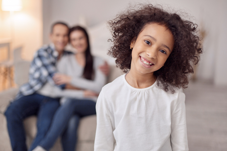 A little girl is smiling in front of a family sitting on a couch.