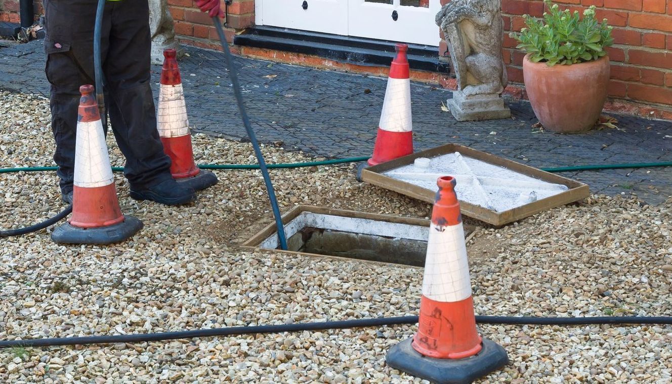 Worker beside orange traffic cones on gravel near a utility hatch and a building wall