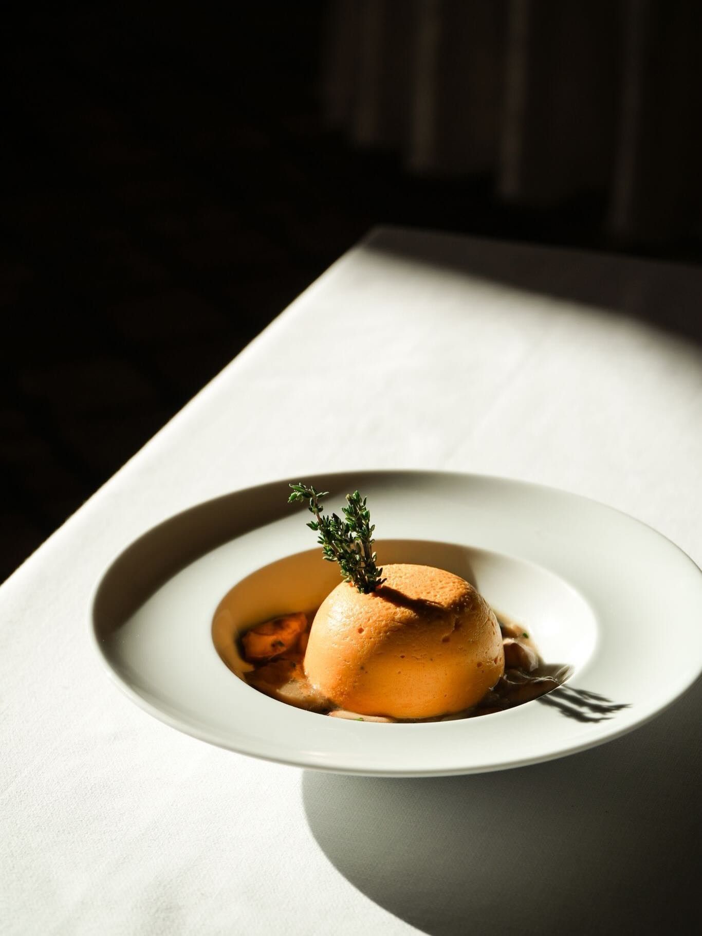 Un dessert elegante su un piatto bianco con salsa e un rametto di erbe aromatiche su un tavolo illuminato dal sole.