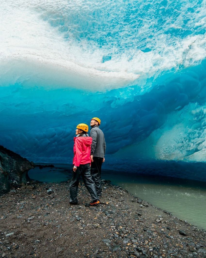 Two hikers in helmets stand under a bright blue ice cave ceiling beside a shallow stream.