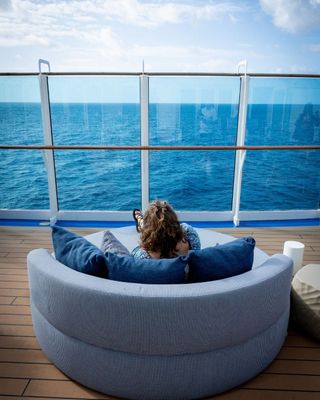 Person relaxing in a round chair on a cruise ship deck, facing the blue ocean and sky.