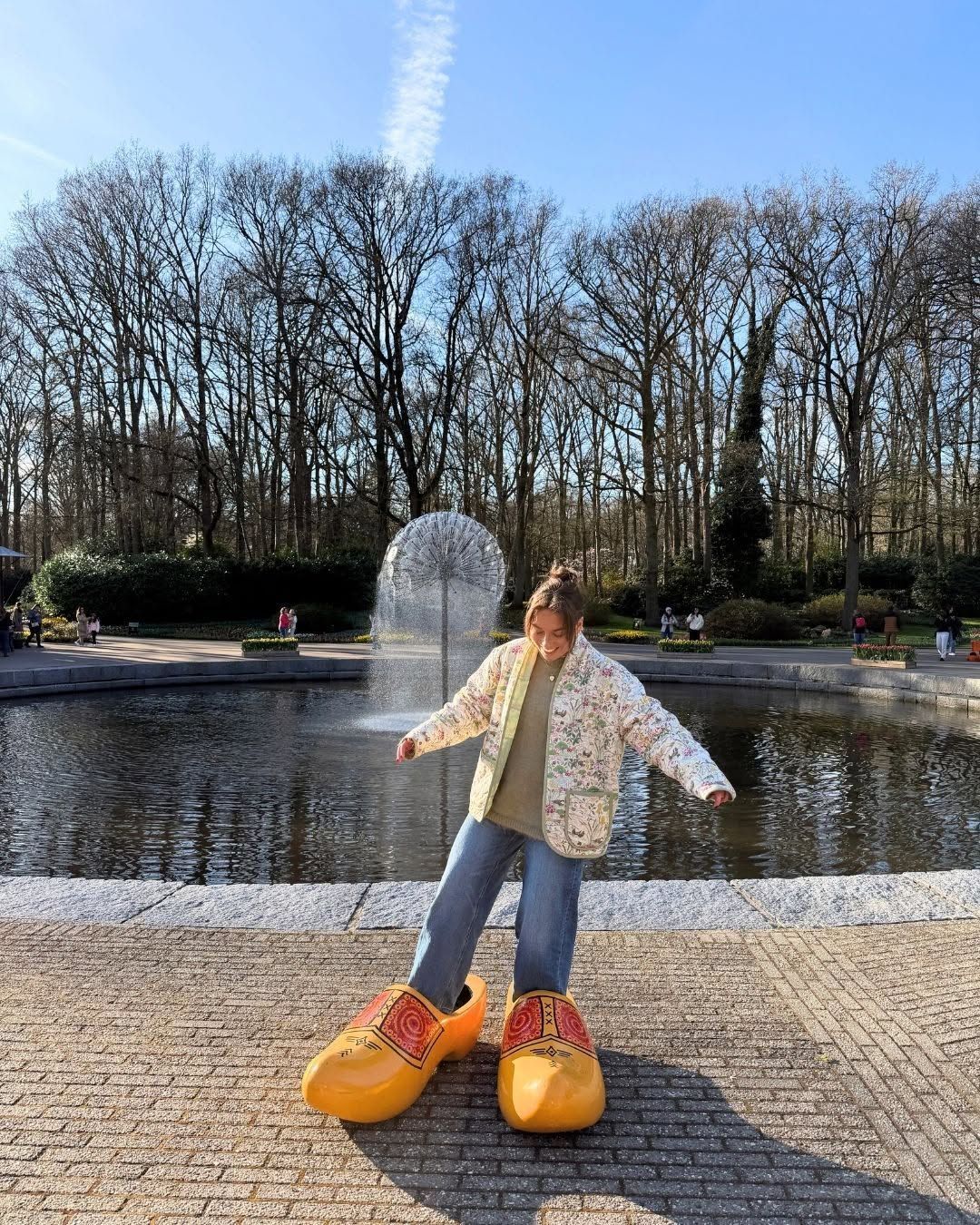 Person standing by a pond in oversized orange shoes, with bare trees and water in the background