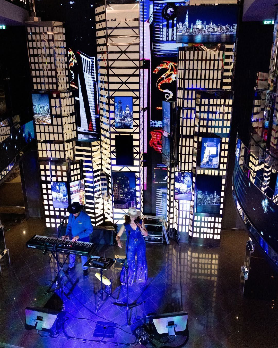 Blue-lit stage with a drum kit and musicians framed by illuminated cityscape panels