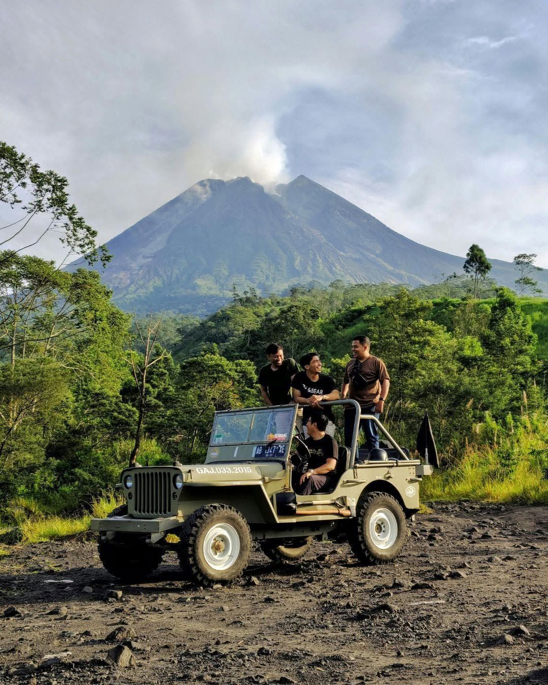 Jeep with people on a muddy track in lush jungle, with a smoking volcano in the background