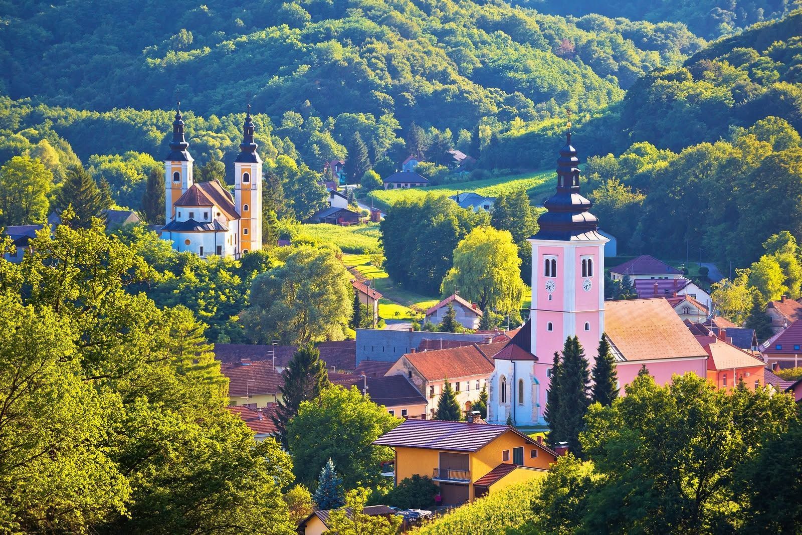 Colorful village with pink church towers nestled among lush green hills and trees