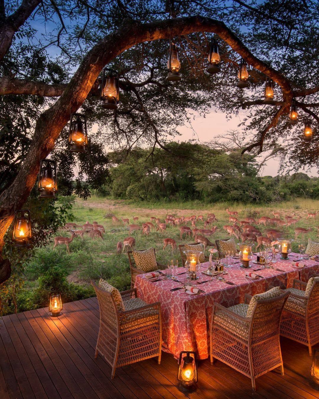 Lantern-lit outdoor dining table beneath a curved arch in a twilight garden garden with flowers and trees