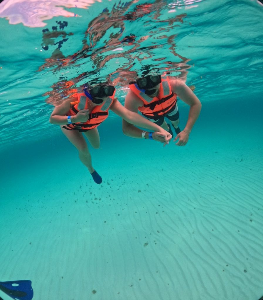 Two snorkelers in orange life vests swimming in clear turquoise water above a sandy seafloor