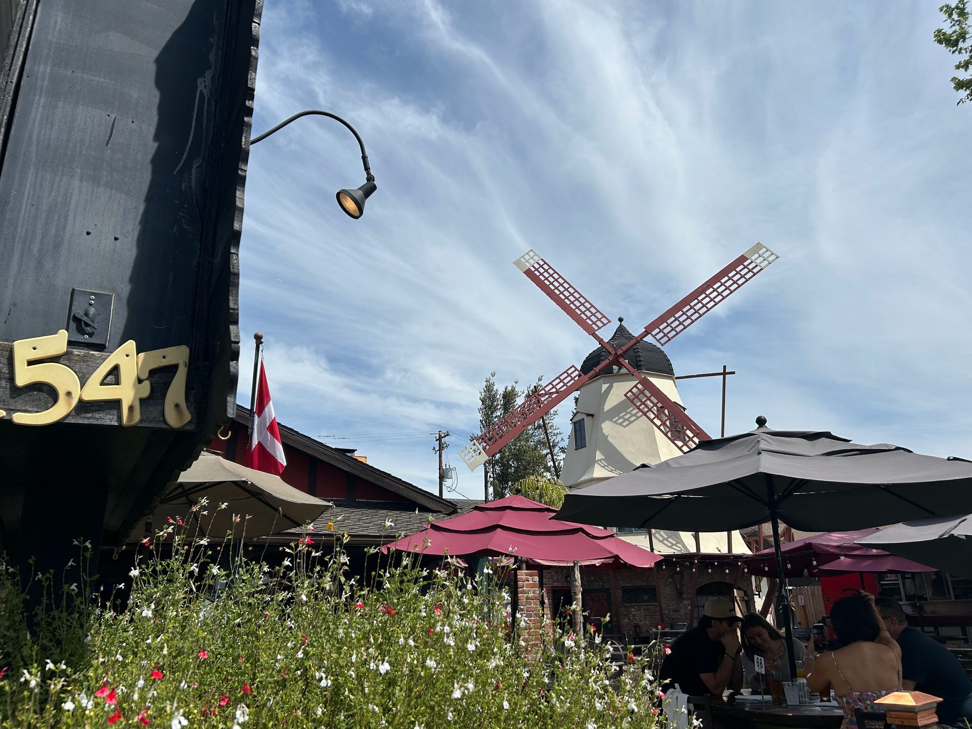 Windmill, patio umbrellas, and flowers under a blue sky at a waterfront cafe.