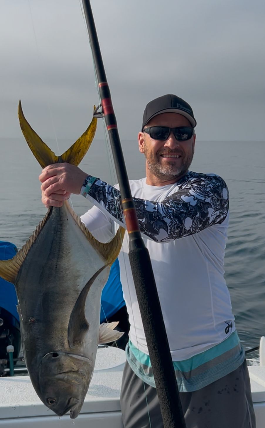 Man holding a large silver fish by the sea, wearing sunglasses and a cap on a boat