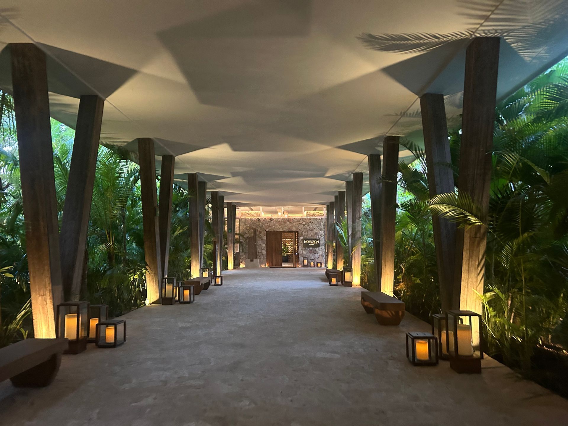 Covered outdoor walkway lit by lanterns, with columns, plants, and a glowing building entrance ahead