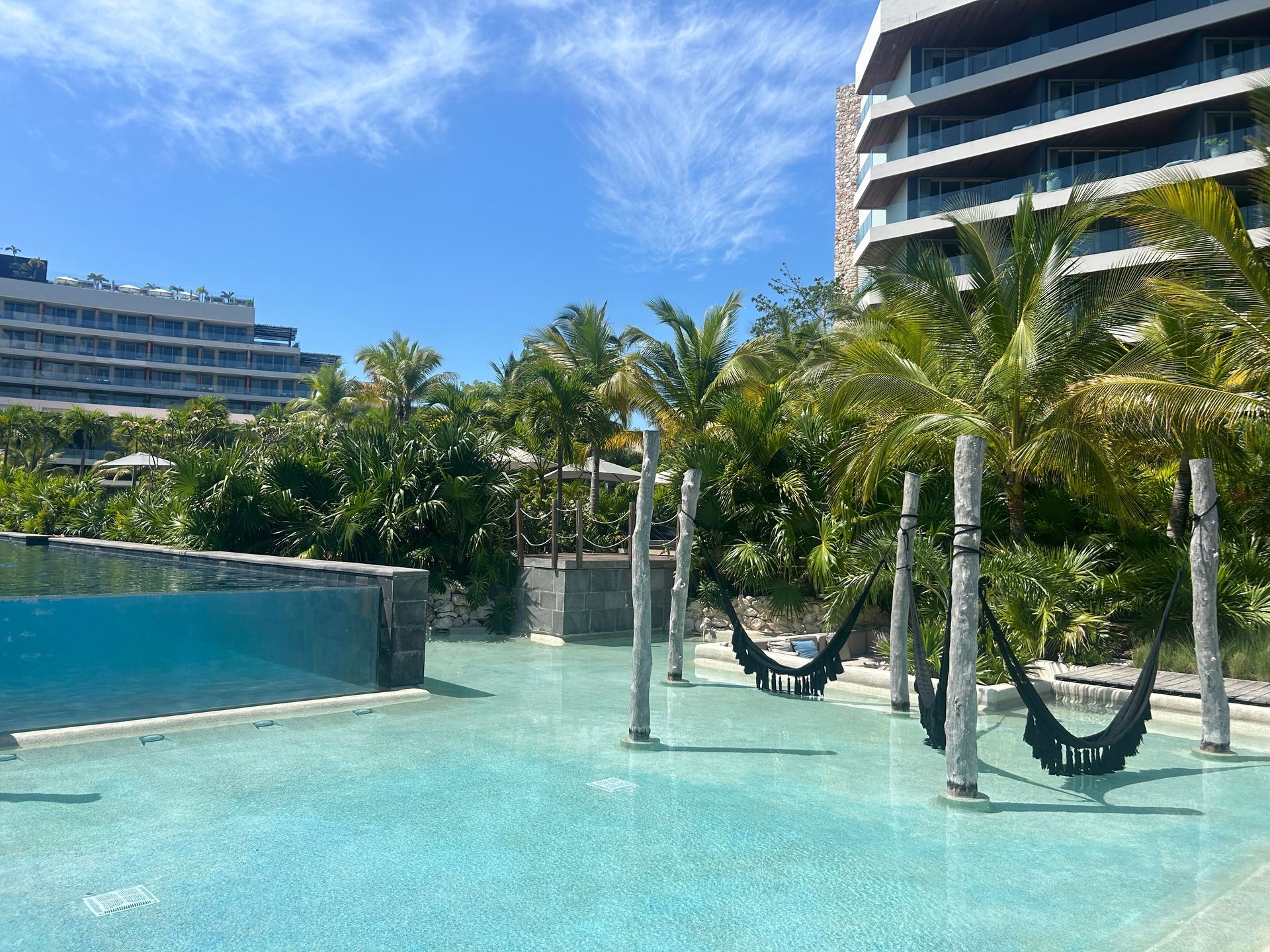 Tropical resort pool with turquoise water, palm trees, hammocks, and oceanfront hotel buildings
