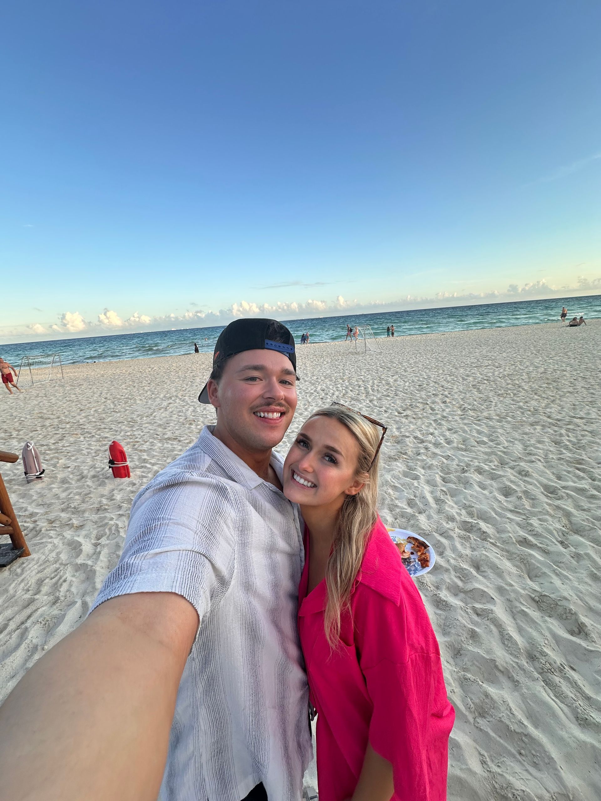 Couple taking a selfie on a sandy beach at sunset, smiling with ocean and shoreline behind them