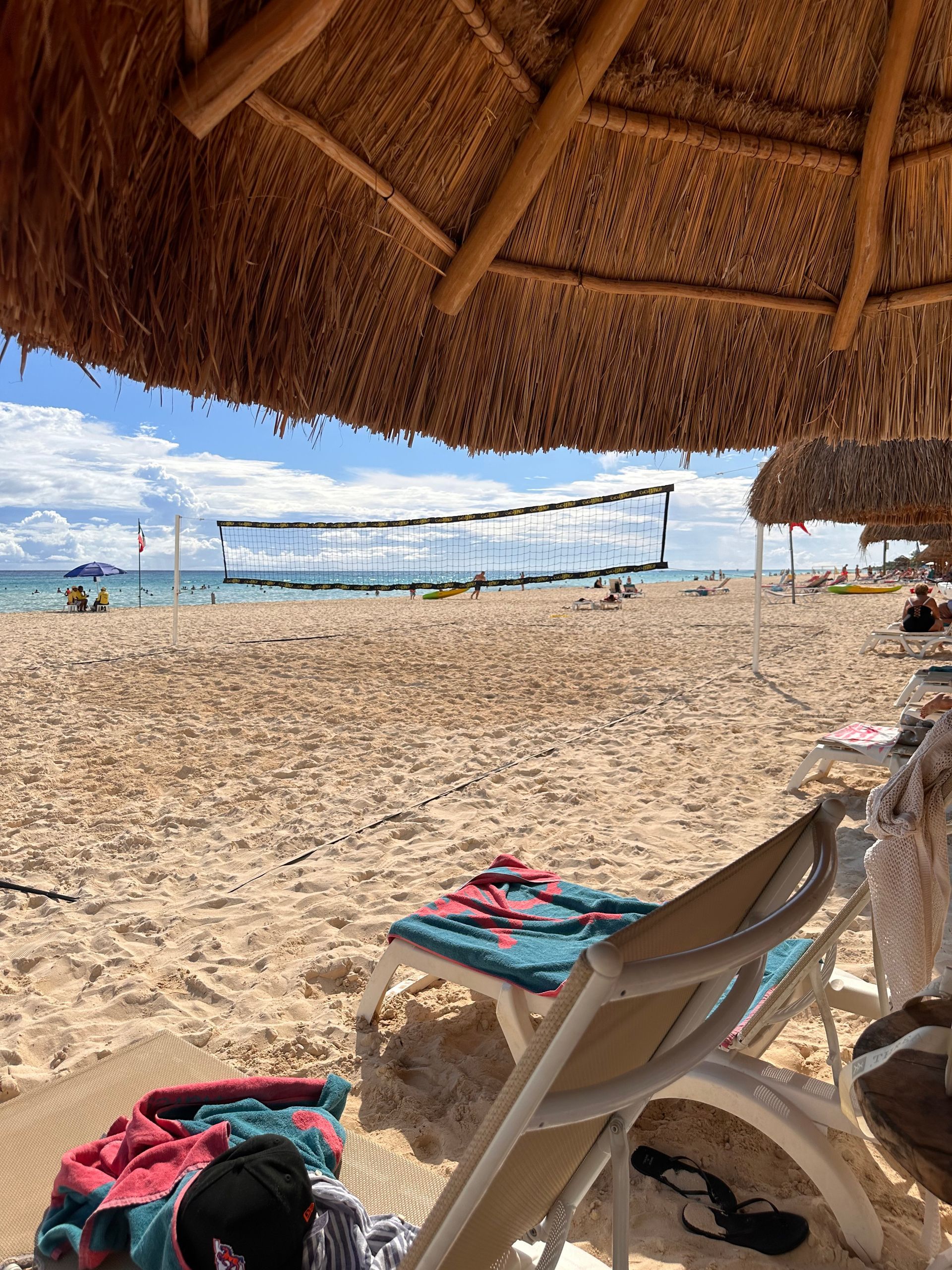 Shaded beach chairs under a thatched umbrella facing a sunny sandy beach and blue ocean