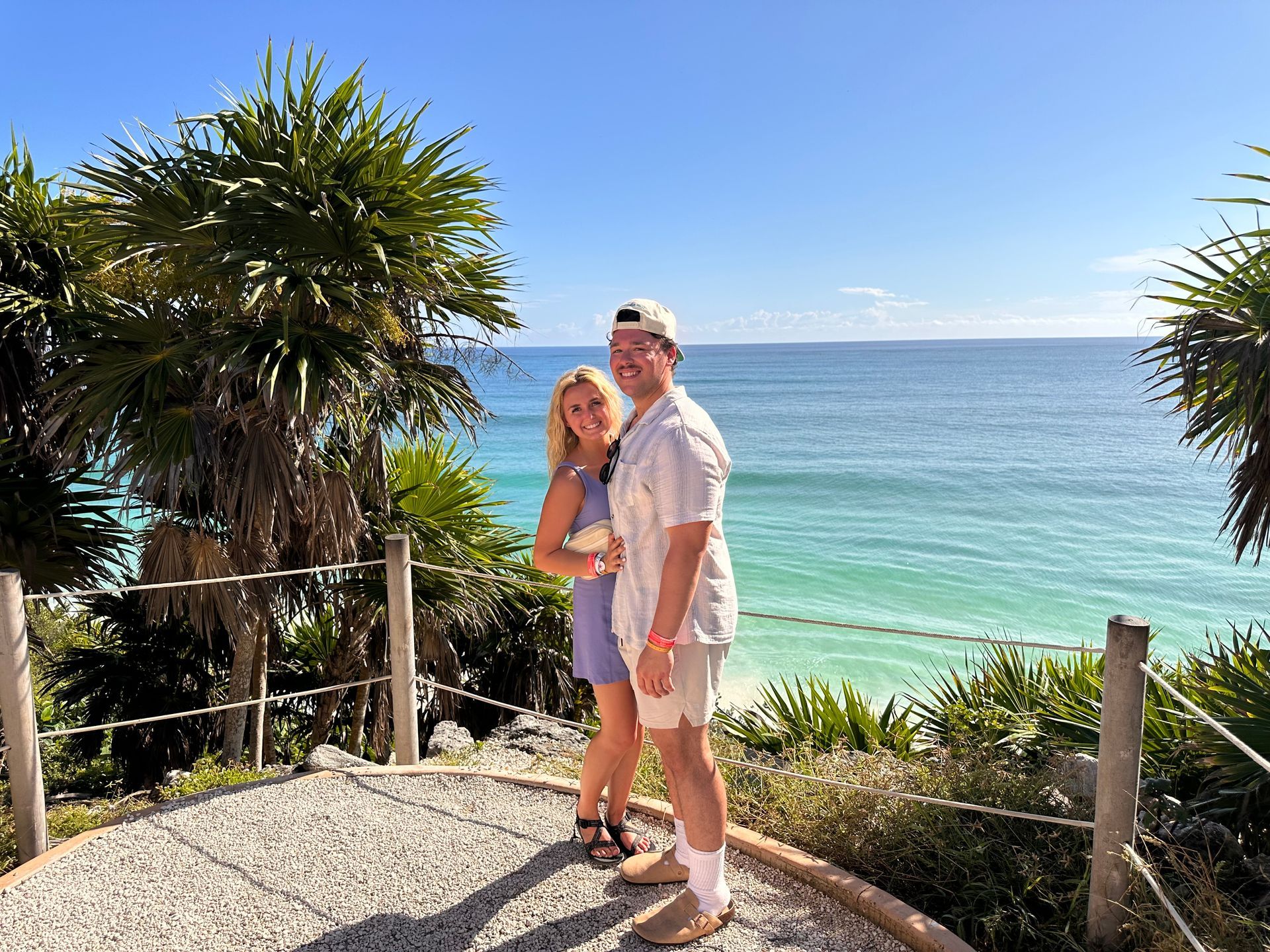 Two people standing on a coastal overlook with palm trees and turquoise ocean in the background