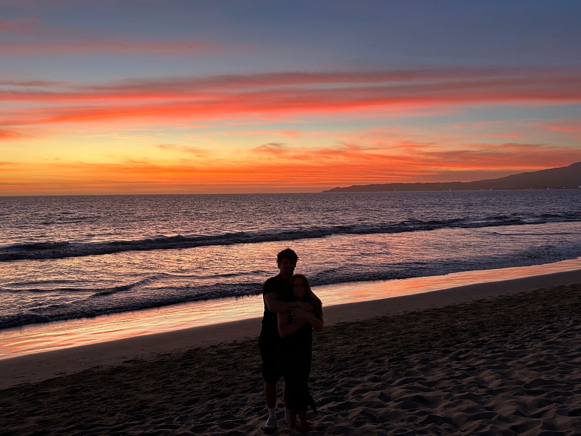 Person standing on a beach at sunset, with orange and pink sky over the ocean.