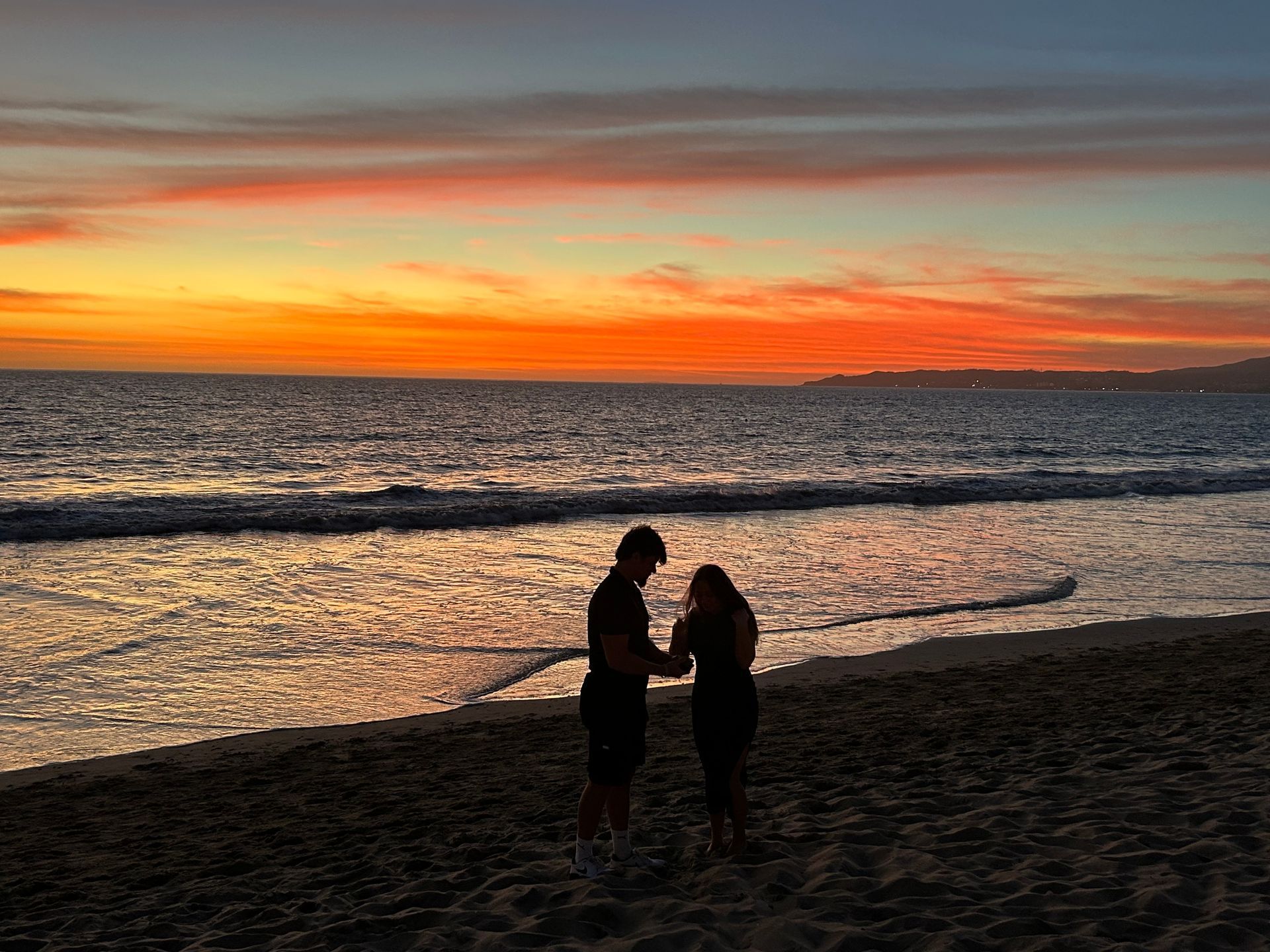 Two people silhouetted on a beach at sunset, with orange sky and waves in the background