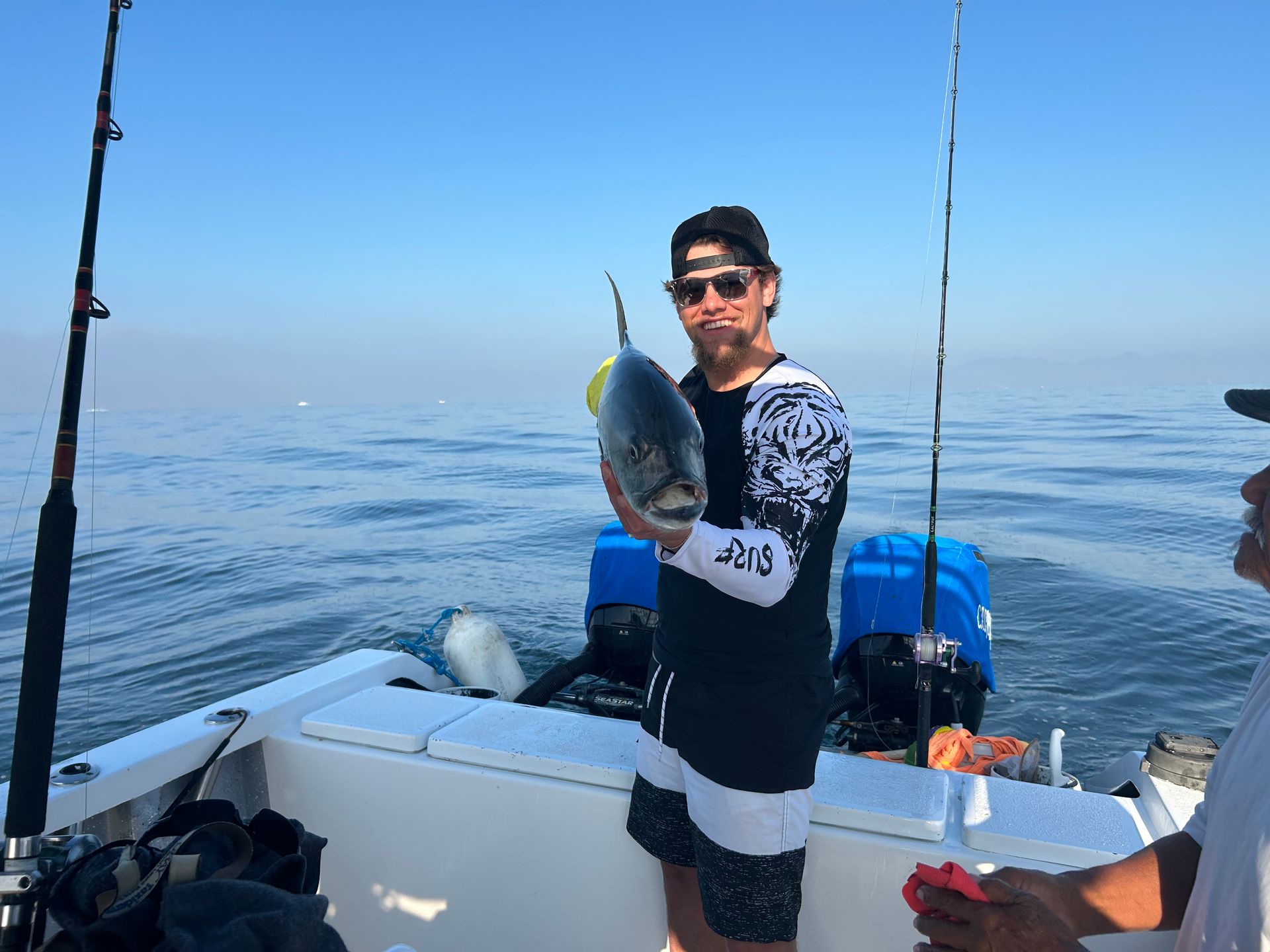 Angler on a boat holding a fish, with rods and calm blue water in the background.