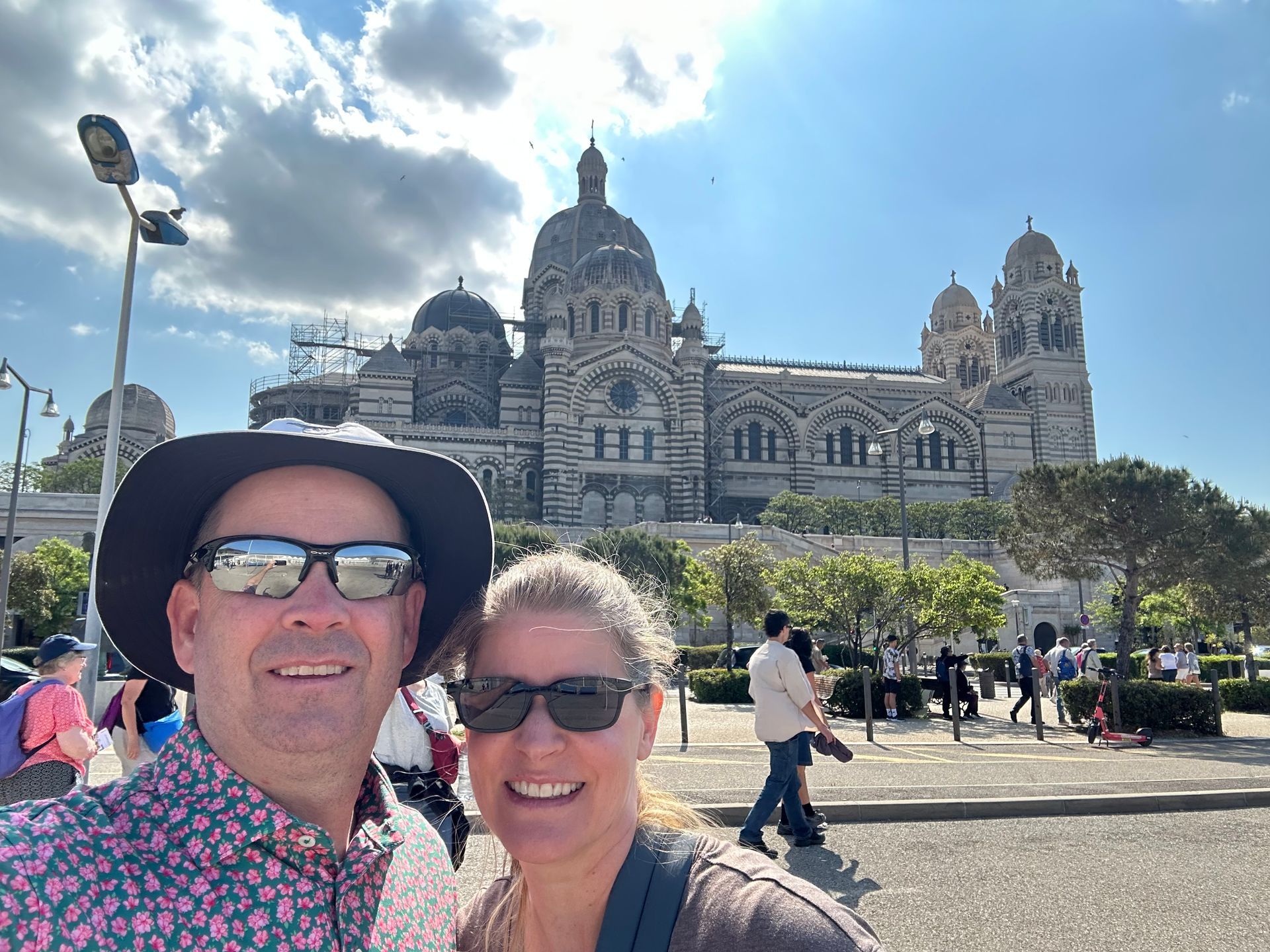 Two smiling people in sunglasses pose in front of a grand historic building under a blue sky