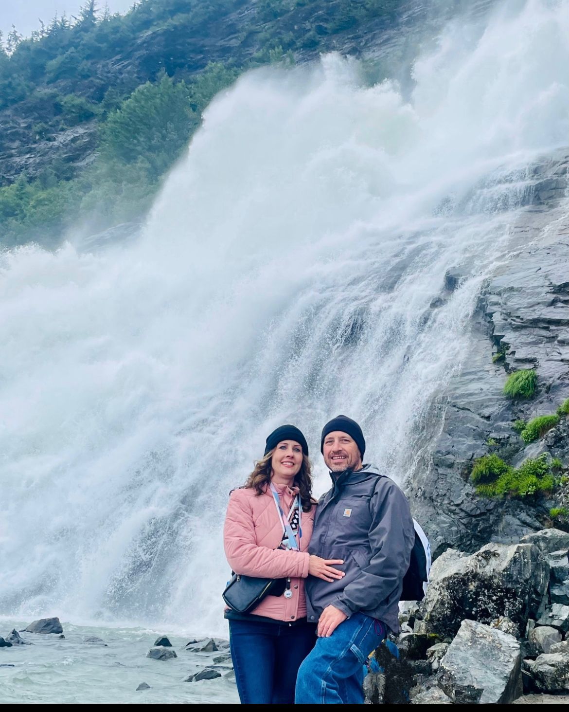 Couple posing in front of a large waterfall, wearing jackets on a rocky shore.