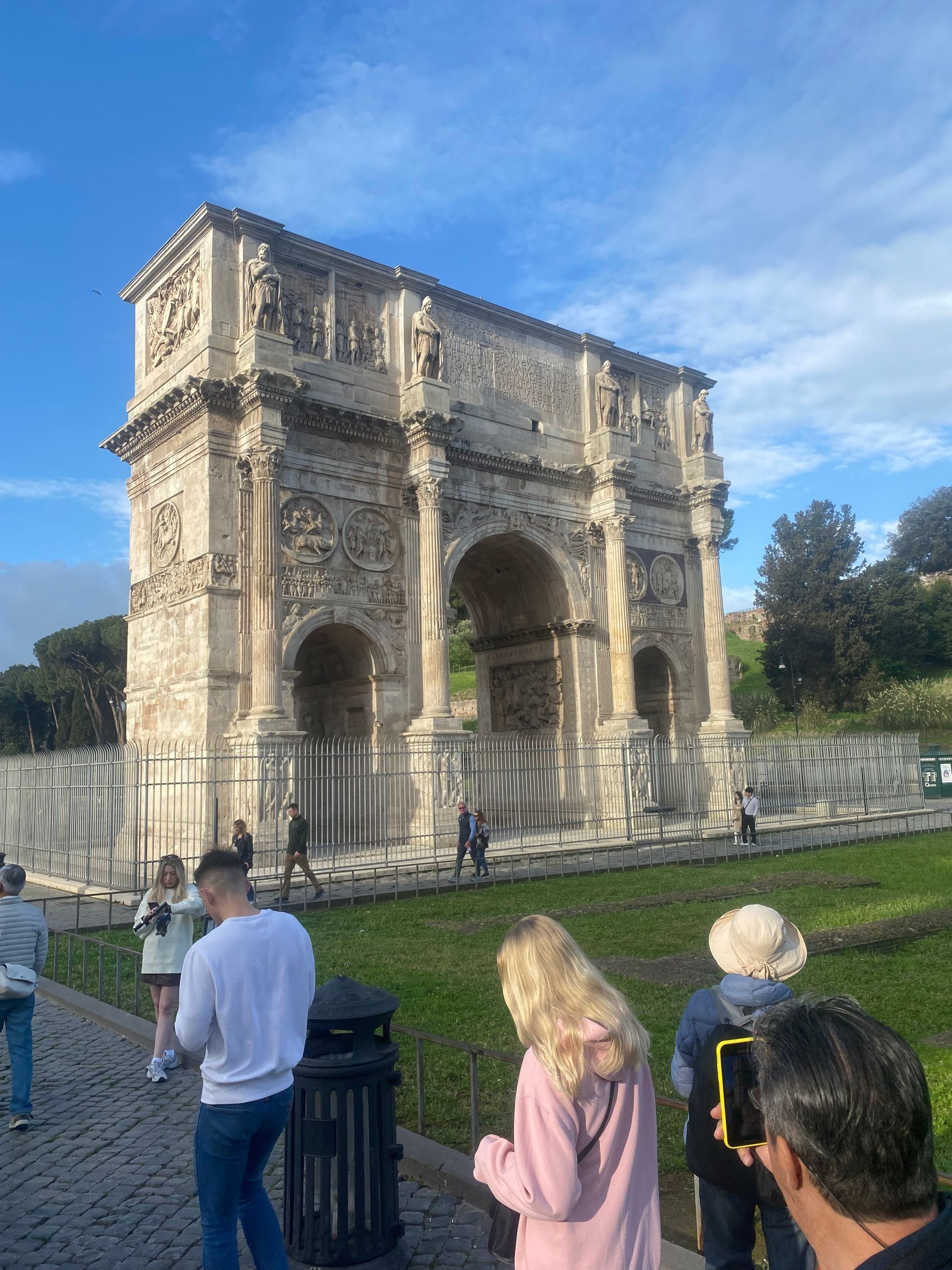 Ancient stone archway in a park, with tourists walking in front under a blue sky.