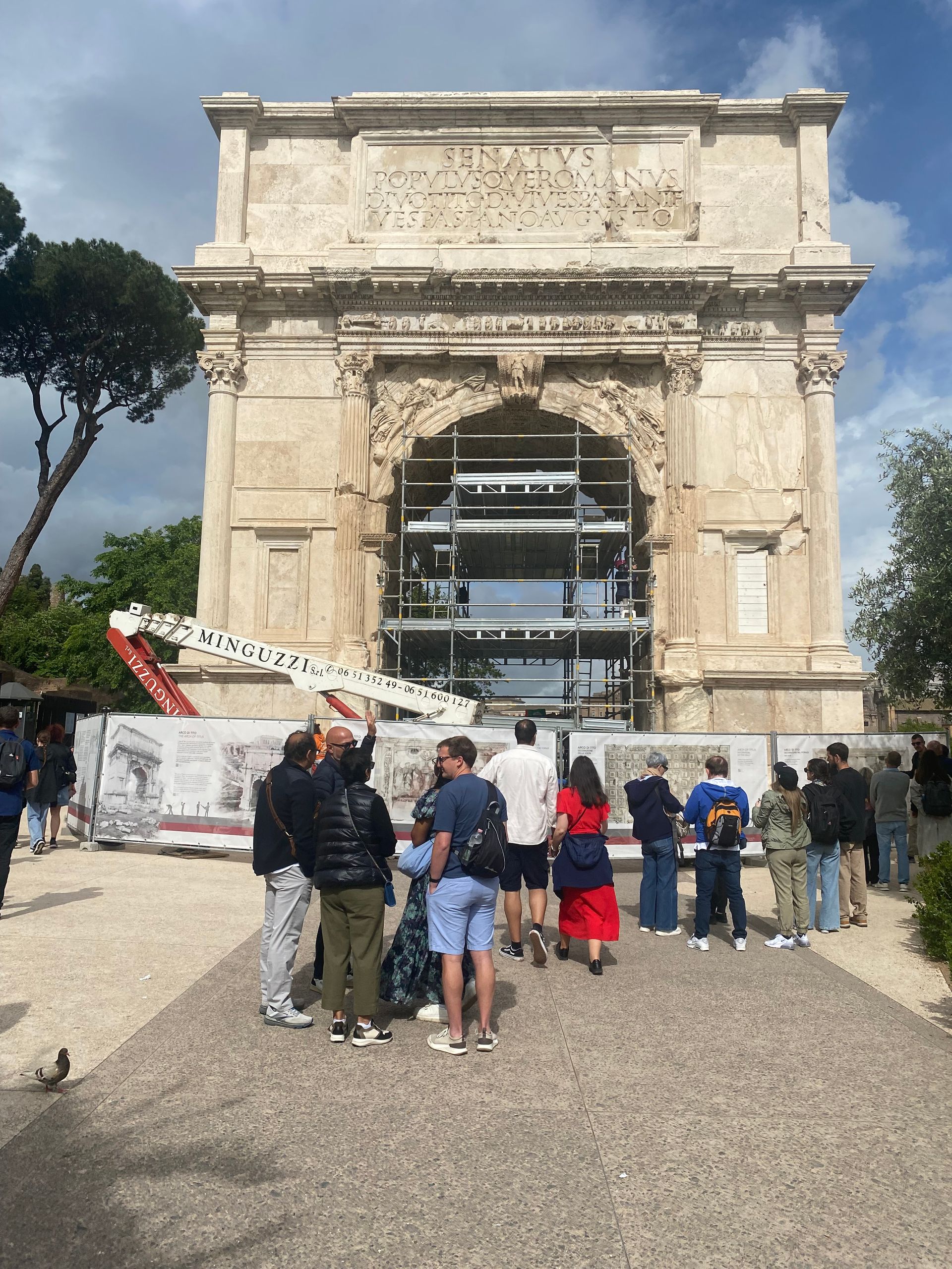 Tourists gathered before a stone arch monument with scaffolding, under a bright sky.