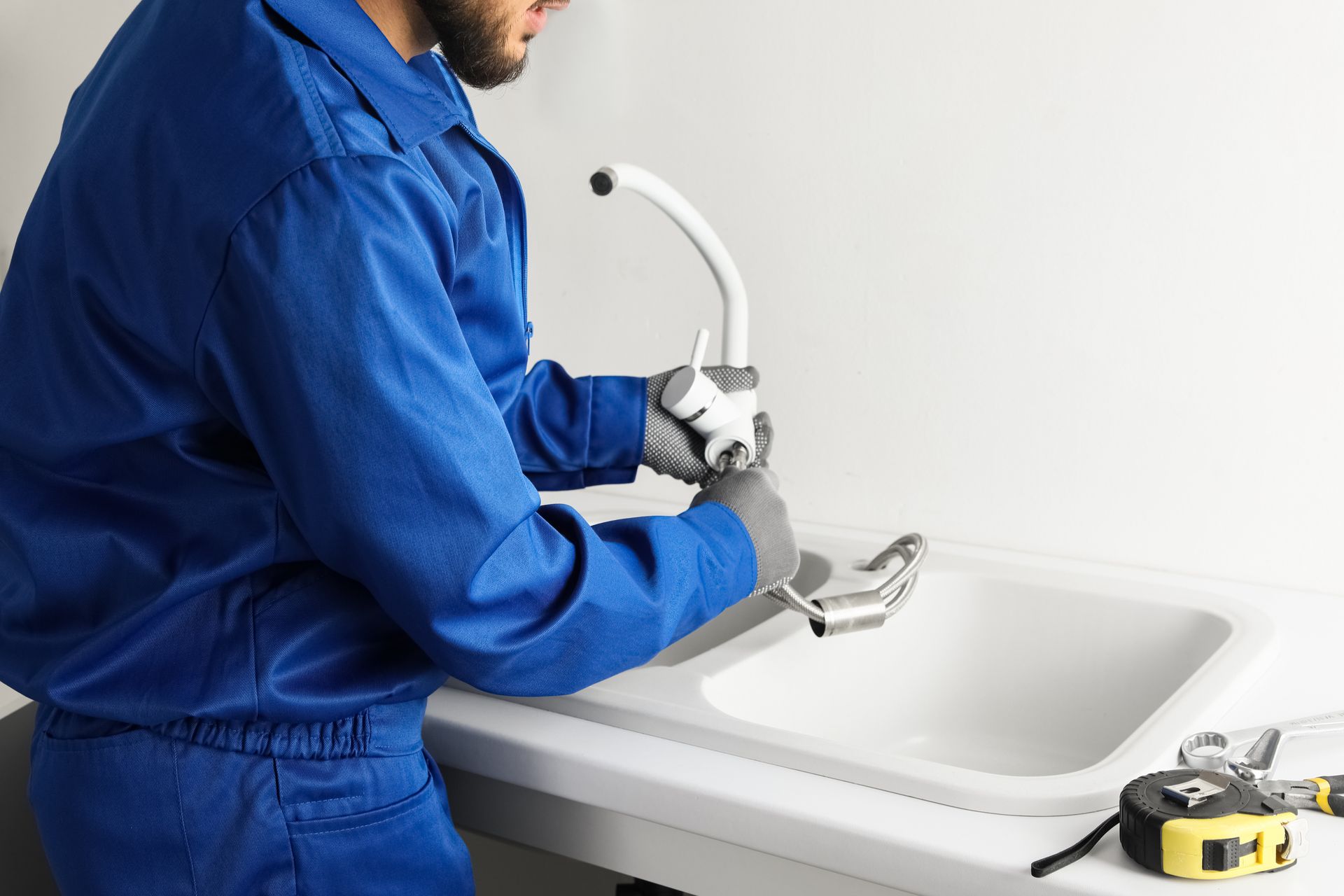 Plumber in blue coveralls fixing a faucet on a white kitchen sink.