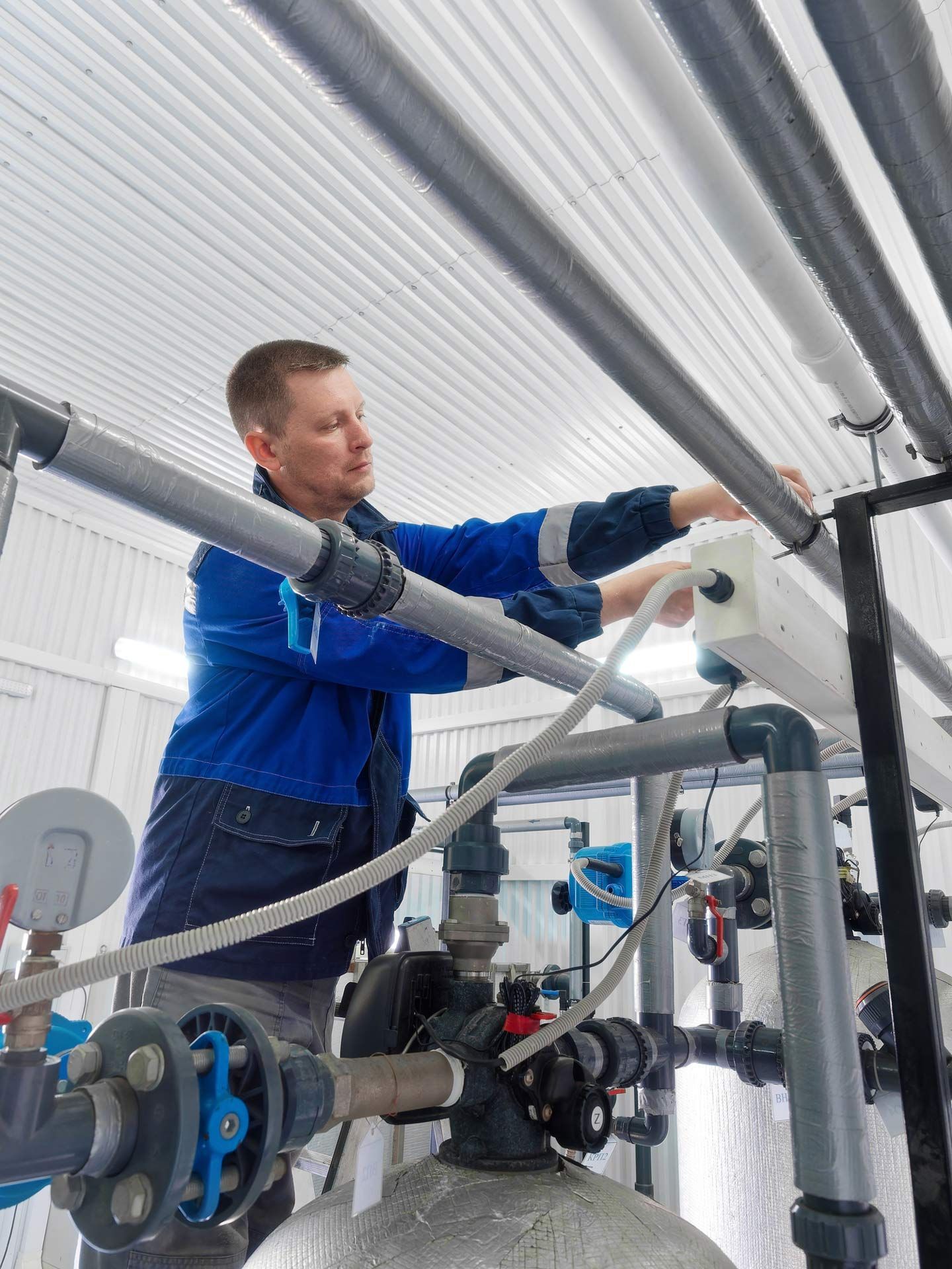 Man in blue jumpsuit works on pipes and equipment inside a white industrial building.