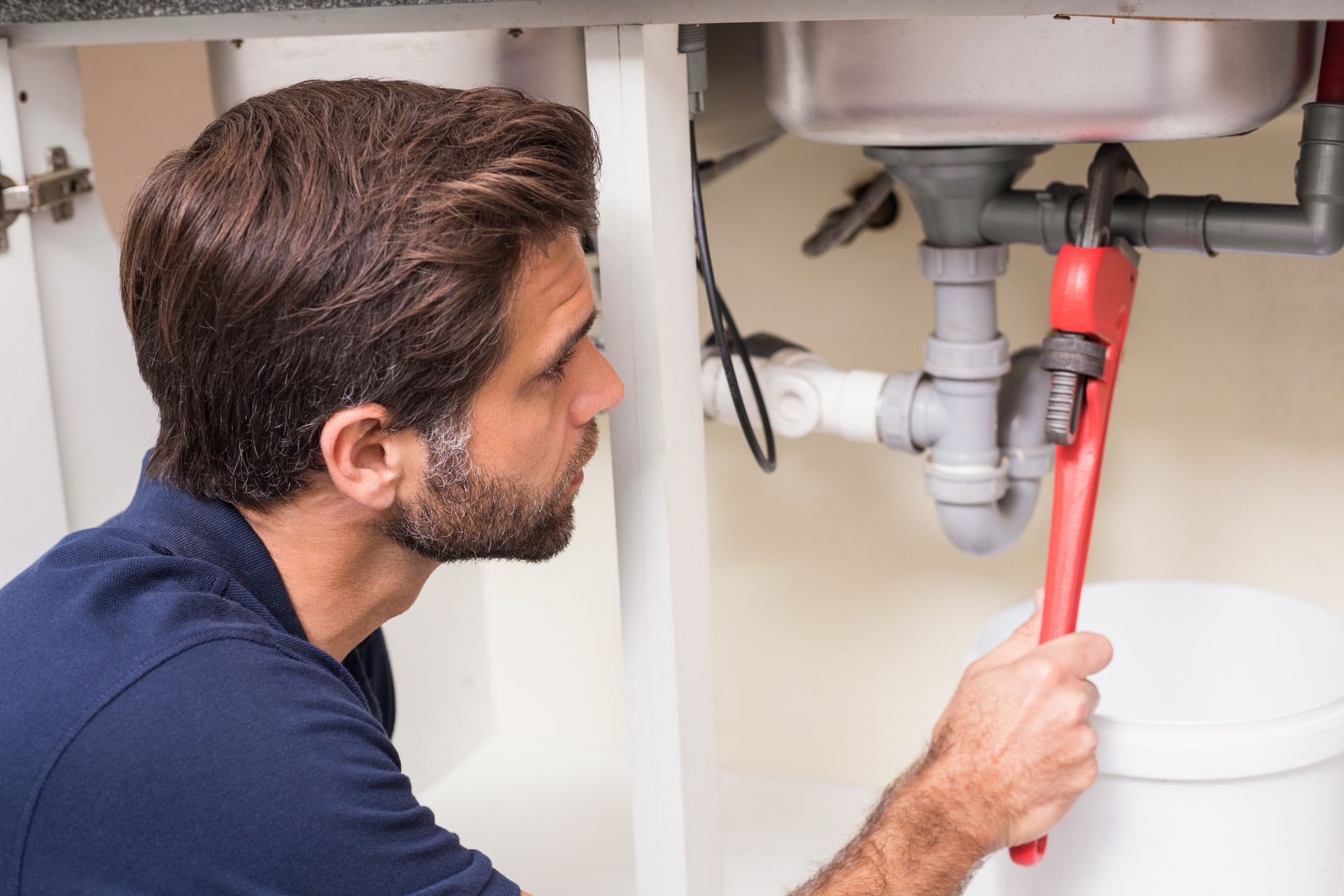 Plumber, red wrench, working under a sink, looking at pipes.