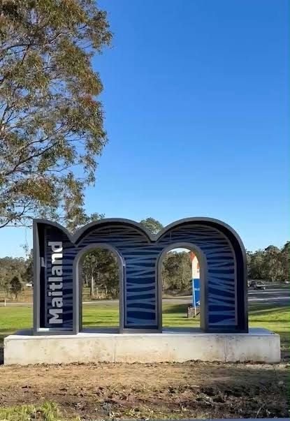 Blue “Maitland” sign sculpture on a roadside under a clear sky, with trees and grass nearby