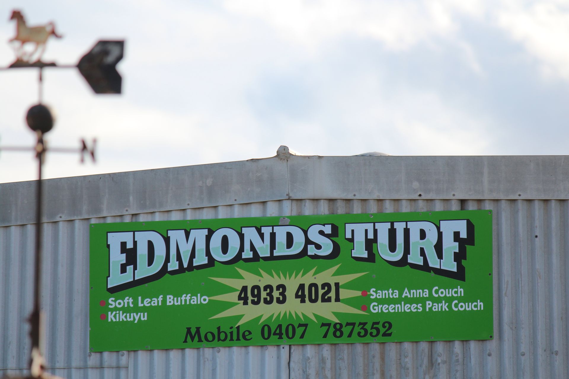 Green sign for EDMONDS TURF on a building, with contact numbers and a weather vane above.