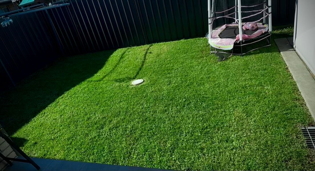 Small backyard with bright green artificial grass, a white pole, and a trampoline by the fence.