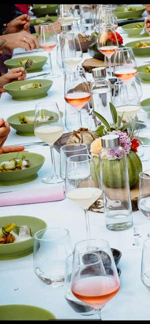 Een lange eettafel gedekt voor een evenement, met groene schalen vol eten, meerdere wijnglazen en bloemstukken in het midden.