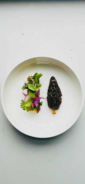Een gerecht op een bord met een kleine salade op een sneetje toast, naast een enkele geroosterde morielje op een wit bord.