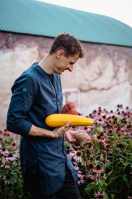 Een persoon in een blauwe tuniek houdt een gele courgette en wortelgroenten vast, staand in een tuin met paarse zonnehoedjes.