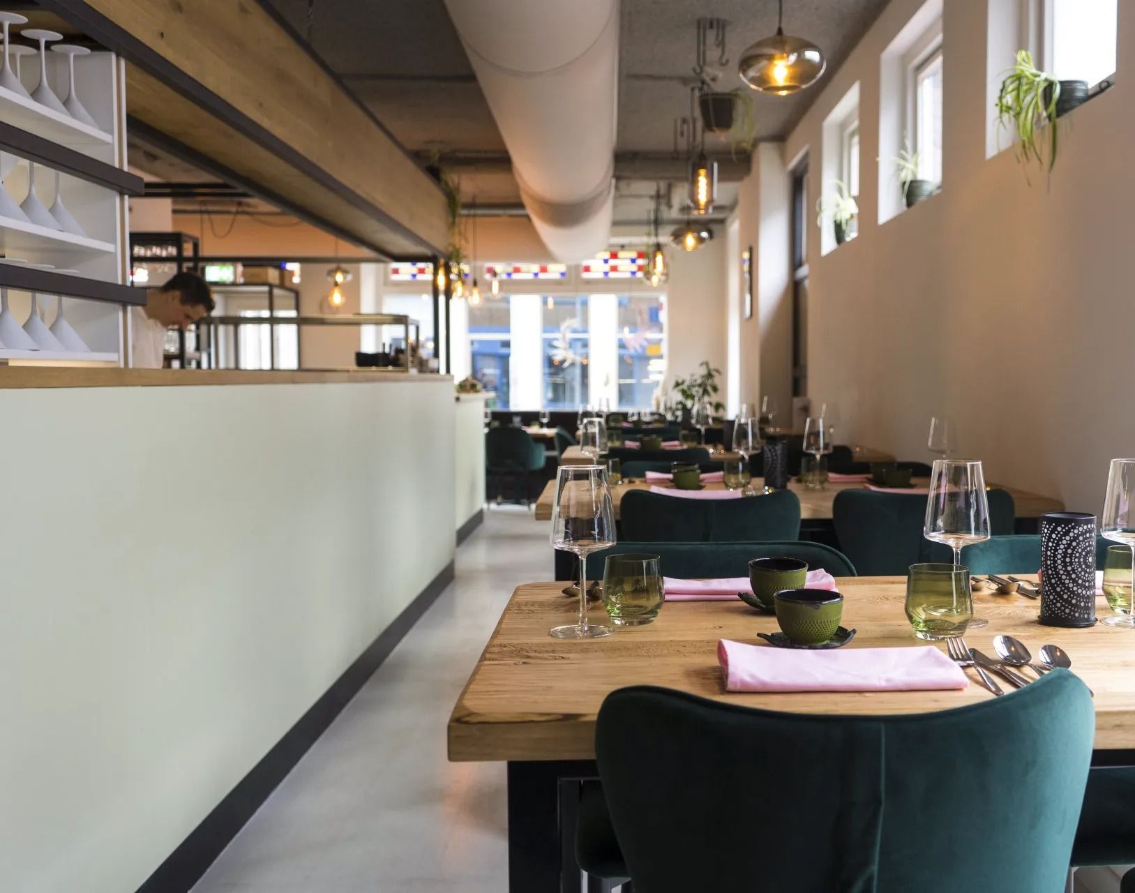 Modern restaurantinterieur met lichte muren, houten tafels en donkergroene gestoffeerde stoelen in een open eetruimte.