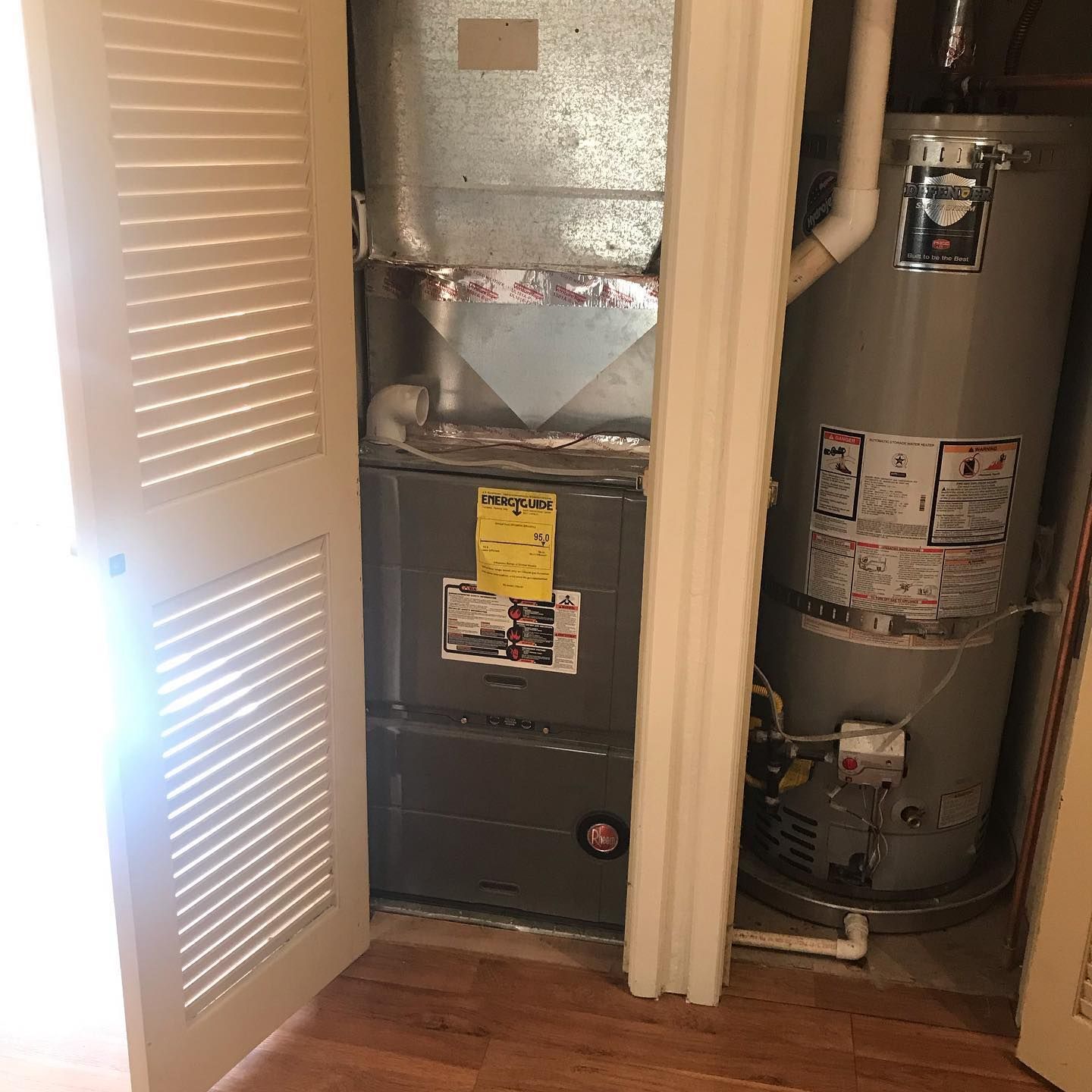 A utility closet with a grey furnace unit and a tall, grey water heater side-by-side on a wooden floor.