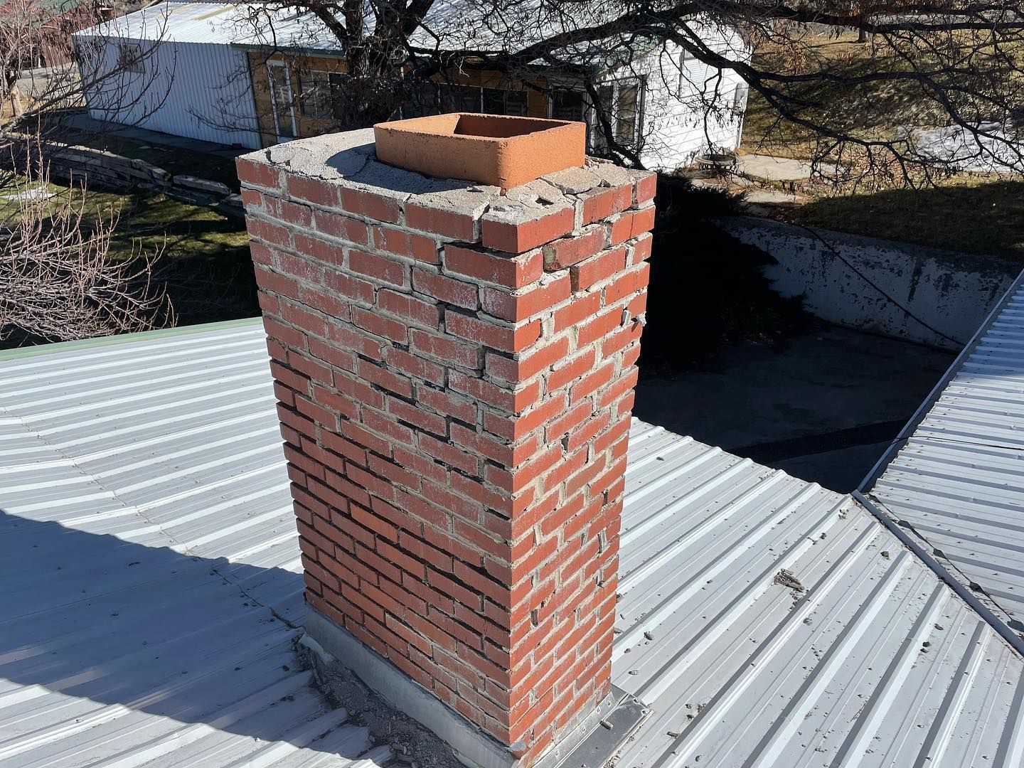 A weathered red brick chimney with a terracotta flue liner stands on a corrugated metal roof in daylight.