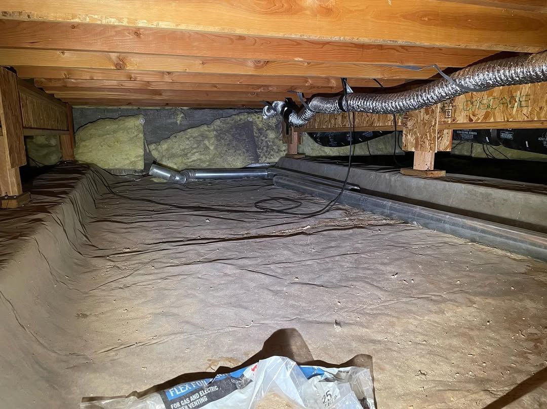 A low, crawl space area showing exposed wooden floor joists, insulation, a vapor barrier on the ground, and flexible ducting.