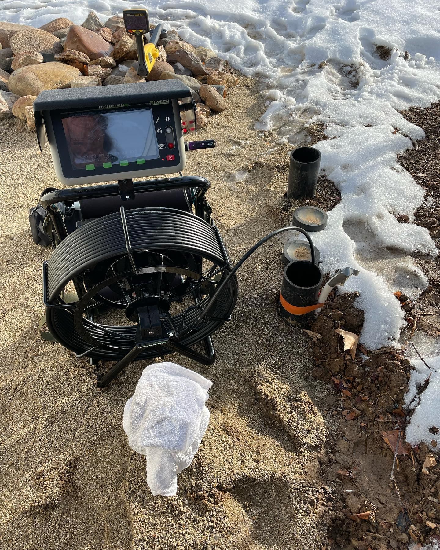 A pipe inspection camera unit with a screen and reel sits on snowy, rocky ground next to an open pipe access point.