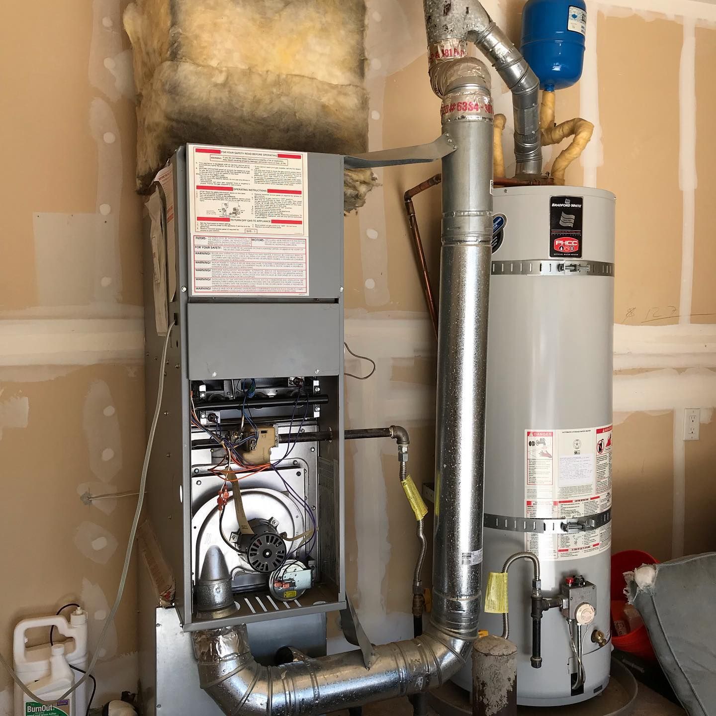 A grey furnace and water heater sit side-by-side against an unfinished wall in a garage, connected by metal vent piping.