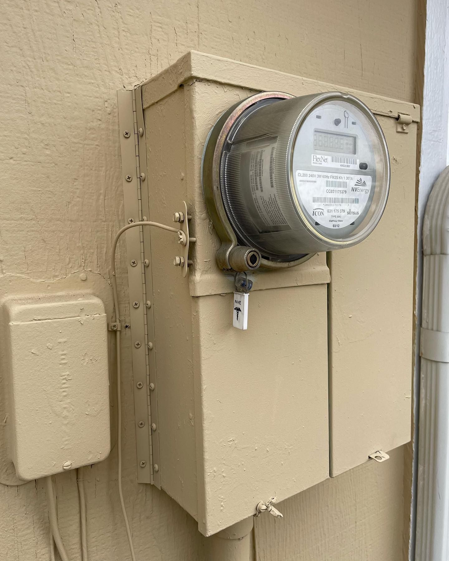 A tan-painted residential electric meter attached to a exterior wall with a small junction box to the left.