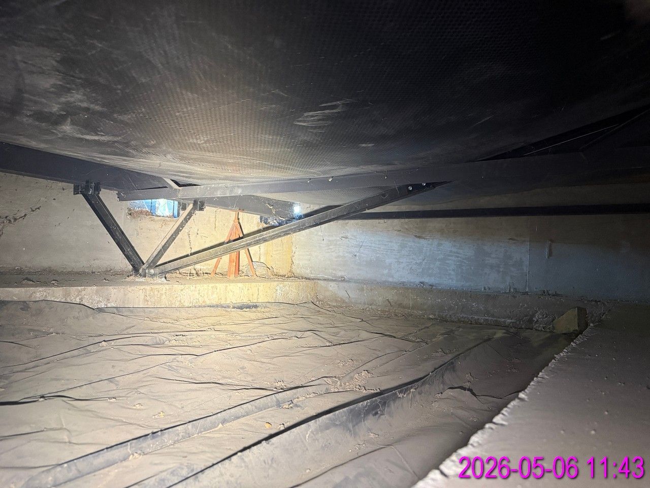 A crawl space with a vapor barrier on the ground, wooden floor joists above, insulation, and a flexible silver duct pipe.