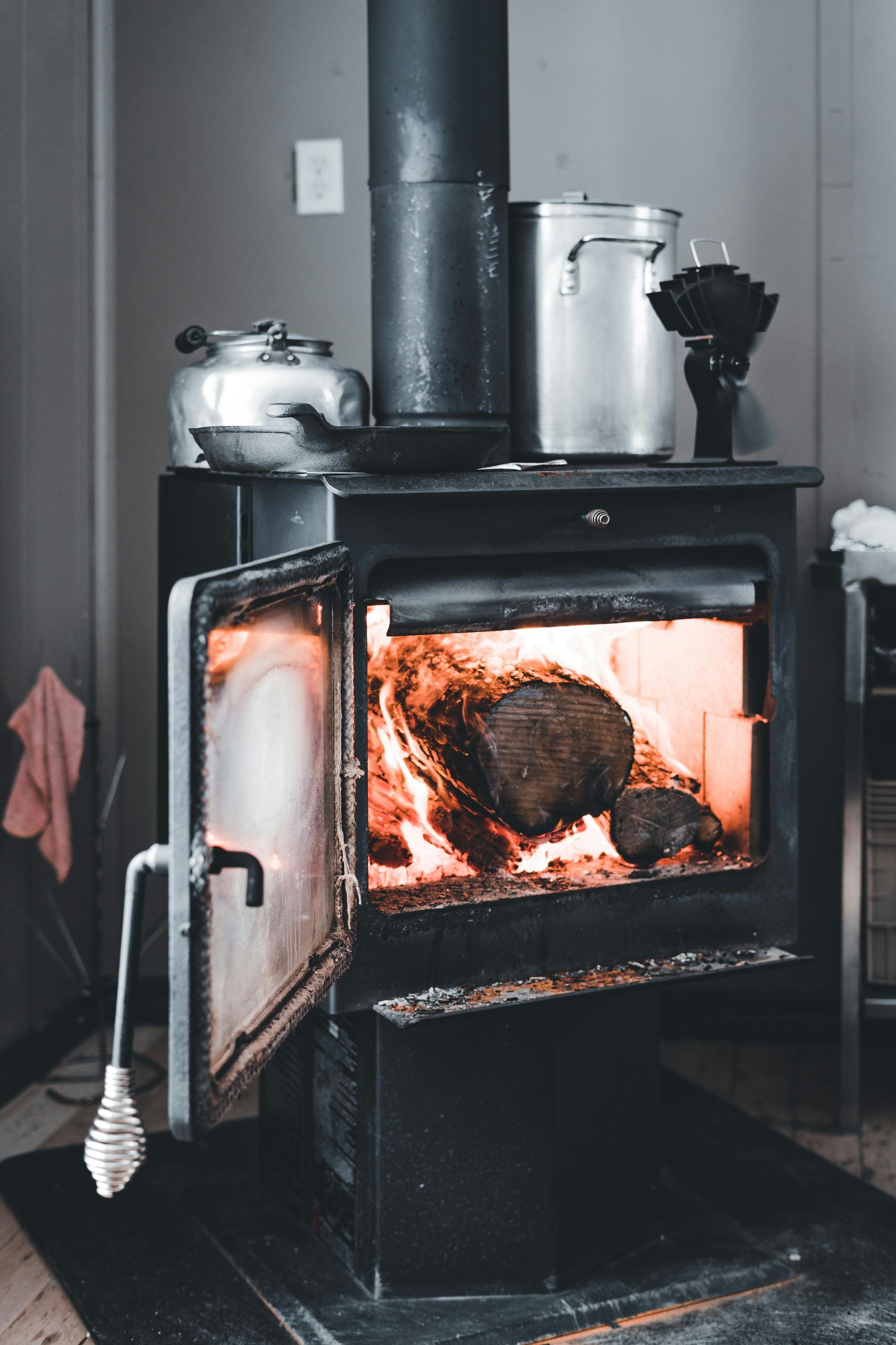 A black wood-burning stove with a glowing fire inside, topped with metal kitchenware, in a gray-walled room.